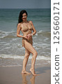 Happy cheerful 51 year old middle aged smiling woman in beige-gold swimsuit bikini standing on the beach near the sea and looking at camera 125660171