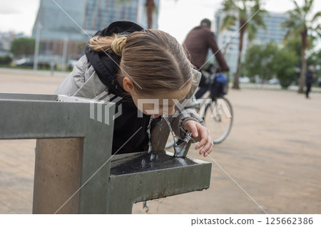 Young girl is drinking water from a fountain. Concept of refreshment and hydration, as the girl Young girl is drinking water from a fountain. Concept of refreshment and hydration, as the girl 125662386