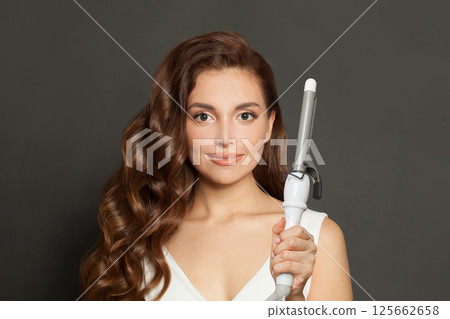Beautiful young model woman with long curly wavy hairstyle showing hot curling tongs iron  125662658