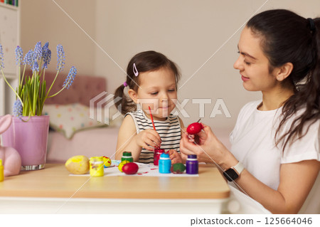 Happy mother and cute little child girl are preparing to celebrate Easter at home. Mom and daughter painting eggs before Easter together. Happy mother and cute little child girl are preparing to celebrate Easter at home. Mom and daughter painting eggs before Easter together. 125664046