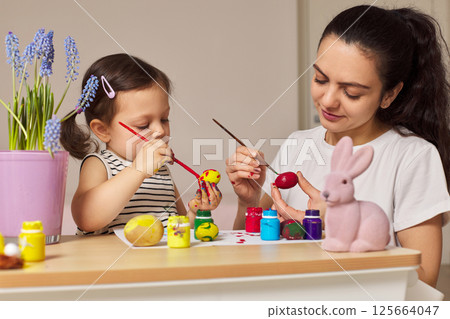 Happy mother teaching cute little child girl to dye and decorate eggs with paints for Easter. Mom and daughter painting eggs before Easter together. Happy mother teaching cute little child girl to dye and decorate eggs with paints for Easter. Mom and daughter painting eggs before Easter together. 125664047