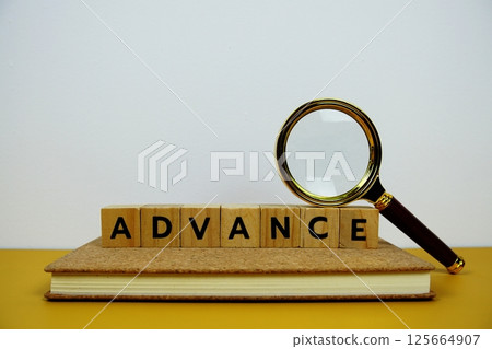 Magnifying glass with wooden blocks alphabet letters ADVANCE text message on yellow background 125664907