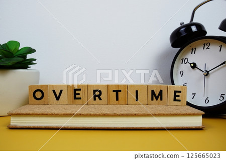 Alarm clock with wooden blocks alphabet letters OVERTIME text message on yellow background 125665023