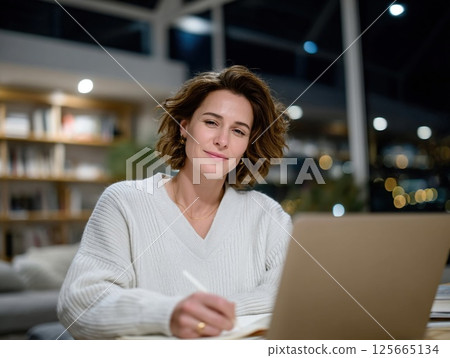 Person is sitting at desk, working on laptop and smiling, in cozy, well lit room with bookshelves in background. atmosphere is relaxed and focused 125665134