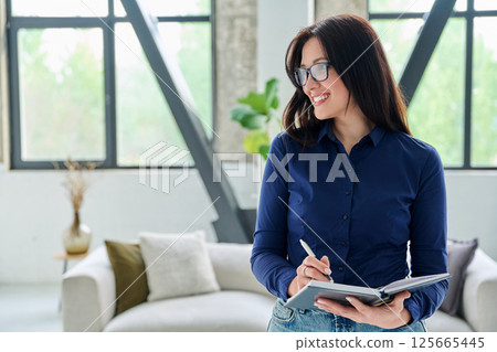 Portrait of smiling business woman with notebook in office with couch Portrait of smiling business woman with notebook in office with couch 125665445
