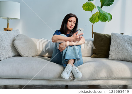 Serious young woman sitting at home on the couch using smartphone Serious young woman sitting at home on the couch using smartphone 125665449