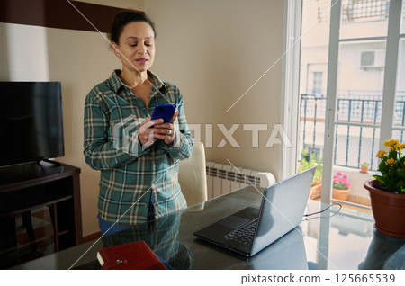 Woman Using Smartphone and Laptop in a Bright Home Office Environment Woman Using Smartphone and Laptop in a Bright Home Office Environment 125665539