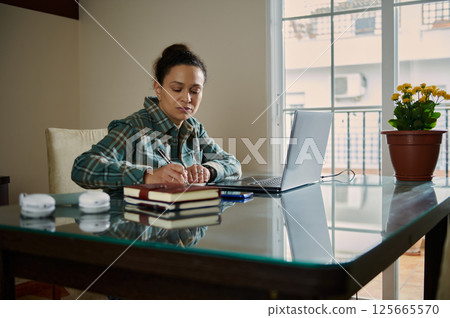 Focused Woman Writing Notes at a Desk with Laptop in Home Office 125665570