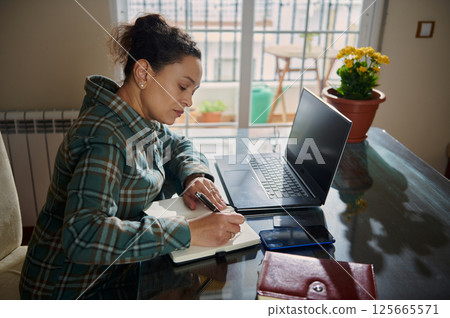 Woman at Desk Working on a Laptop and Taking Notes at Home Woman at Desk Working on a Laptop and Taking Notes at Home 125665571
