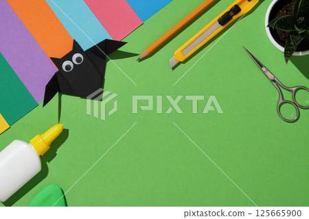 Creative workstation with colorful craft materials on green background with a handmade black origami bat with scissors, utility knife, glue, eraser, pencil and colorful paper sheets. Copyspace 125665900