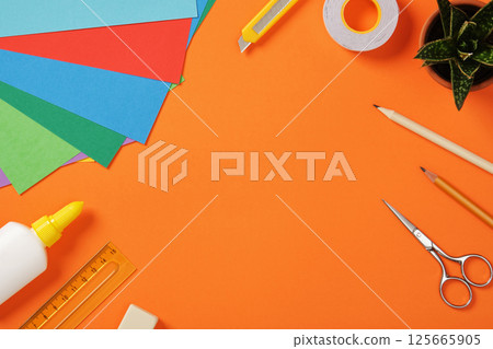 Supplies for paper art hobby: scissors, craft knife, pencil, eraser, colored paper on orange background. Space for your text. Paper crafts work table flatlay. 125665905