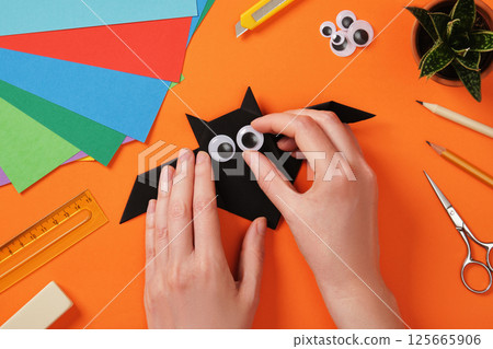 Making an origami bat paper craft. Women's hands attach an eye to a paper toy. Hobby idea. 125665906