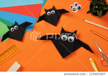 Three handmade black origami bats with googly eyes on a bright orange background surrounded by colorful paper sheets and crafting tools 125665907
