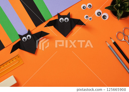 Origami paper bats with goggle eyes on a orange background. Surrounded by crafting supplies, paper sheets, pencil, eraser, glue and knife for DIY projects. Space for text. 125665908