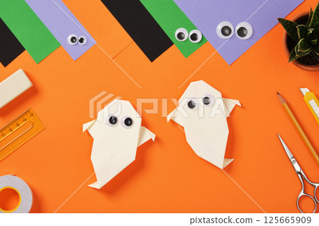 Origami paper ghosts with googly eyes surrounded by colorful craft supplies on orange background. Paper sheets, scissors, markers, glue and knife for kid's crafts or holiday DIY projects. 125665909