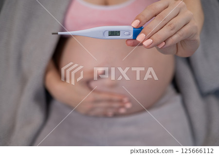Pregnant woman measures temperature while sitting on the couch. Close up of the belly. 125666112