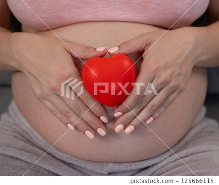 Caucasian pregnant woman holding heart on belly background. Third trimester. Caucasian pregnant woman holding heart on belly background. Third trimester. 125666115