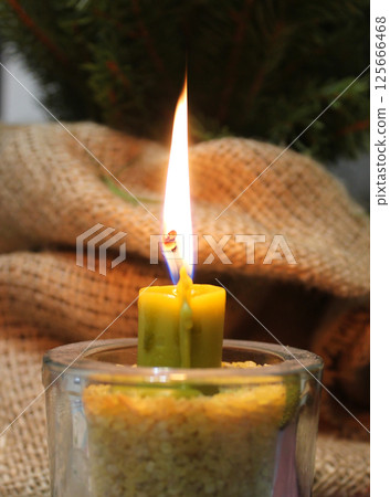 Fire Of Dark Yellow Wax Candle In Glass Candlestick Filled With Grains Near Handmade Burlap Wrapping 125666468