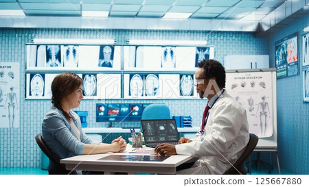 Expert evaluating diagnostic results with a patient in a medical cabinet, showcasing advanced healthcare technology using a screen displaying x rays and MRI scans. Care management. Camera B. 125667880