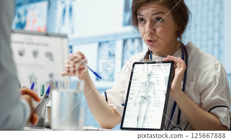 Healthcare provider presenting lab test results to a patient in cabinet, reviewing x ray and MRI scans to assist man through recovery. Medical technology in diagnostics and care management. Camera A. 125667888