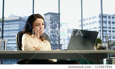Young entrepreneur listening to a podcast while she solves daily business tasks in high end office, corporate success story. Female director working with headphones, determination. Camera A. Young entrepreneur listening to a podcast while she solves daily business tasks in high end office, corporate success story. Female director working with headphones, determination. Camera A. 125667898