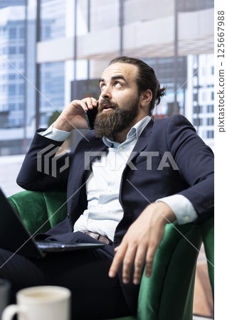 Business administrator speaks with a secretary fixing a meeting on schedule with associates, multitasking on his laptop. CEO busy on a phone call in his corporate office. Communication. Business administrator speaks with a secretary fixing a meeting on schedule with associates, multitasking on his laptop. CEO busy on a phone call in his corporate office. Communication. 125667988