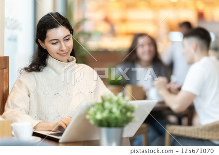 In cozy cafe hall, freelance girl works with laptop and drinks coffee. In cozy cafe hall, freelance girl works with laptop and drinks coffee. 125671070