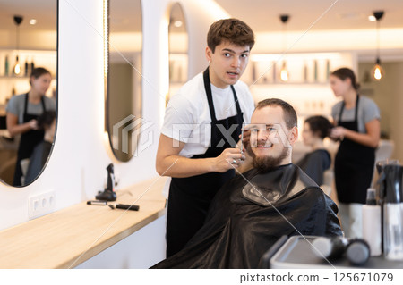 Young man barbershop employee stands next to male client, discussing details of haircut Young man barbershop employee stands next to male client, discussing details of haircut 125671079