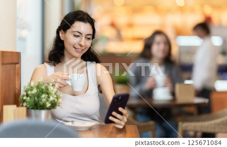 Woman holding mobile phone in her hands and drinking coffee while sitting at a table 125671084