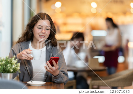 Woman holding mobile phone in her hands and drinking coffee while sitting at a table Woman holding mobile phone in her hands and drinking coffee while sitting at a table 125671085