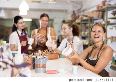 Young woman sits near table and decorates clay ceramic craft vase, plate 125671141