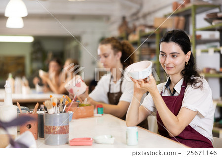 Young woman sits near table and decorates clay ceramic craft vase, plate 125671146