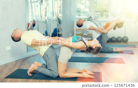 Lovers of active lifestyle perform pair yoga supported backbend on knee in gym Lovers of active lifestyle perform pair yoga supported backbend on knee in gym 125671436