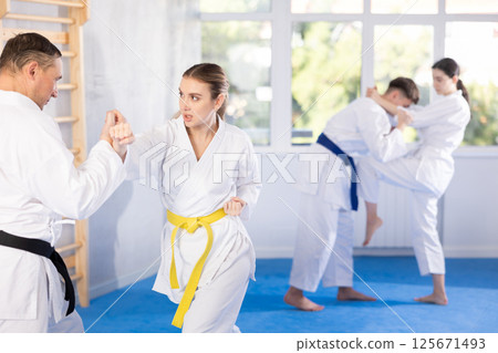 Young female attendee of karate classes fighting with middle-aged man in sports hall Young female attendee of karate classes fighting with middle-aged man in sports hall 125671493