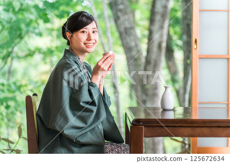 一位身穿浴衣的年輕亞洲女子在旅館客房的門廊上放鬆地喝著清酒。 125672634