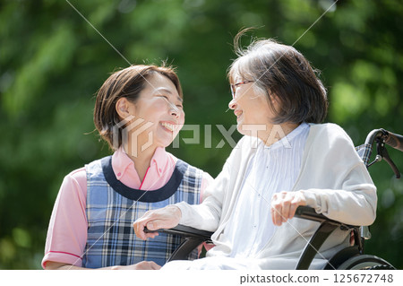 Close-up of a caregiver and an elderly person looking at each other with smiles on their faces as they take a walk in the greenery 125672748