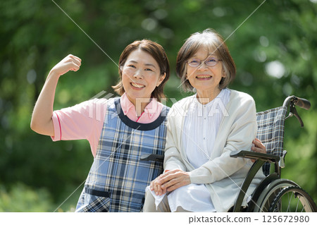 A close-up of a caregiver and an elderly person walking in the greenery, looking at the camera and smiling. 125672980
