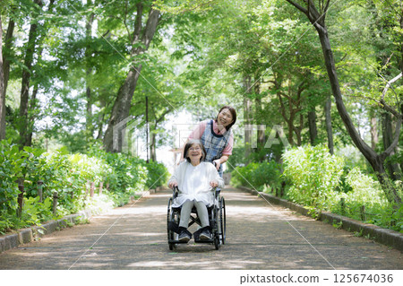 Full body of a caregiver and an elderly person pushing a wheelchair in the fresh greenery Full body of a caregiver and an elderly person pushing a wheelchair in the fresh greenery 125674036