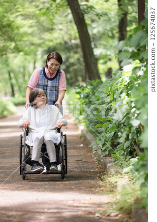 Full body of a caregiver and an elderly person walking while pushing a wheelchair in the fresh greenery 125674037