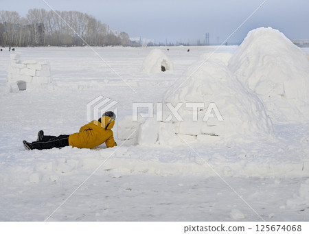 Boy lay down next to the igloo, a national shelter from the cold of the northern peoples, in winter 125674068