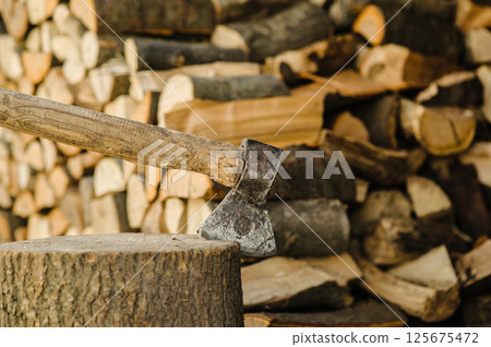 A sharp axe is embedded in a sturdy log. In the background, there are stacked firewood pieces. A sharp axe is embedded in a sturdy log. In the background, there are stacked firewood pieces. 125675472