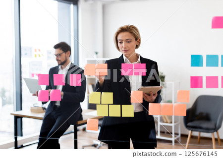 Businesswoman and businessman working collaboratively on project ideas, using post-it notes on transparent glass board. Depicts professional teamwork, corporate planning Businesswoman and businessman working collaboratively on project ideas, using post-it notes on transparent glass board. Depicts professional teamwork, corporate planning 125676145