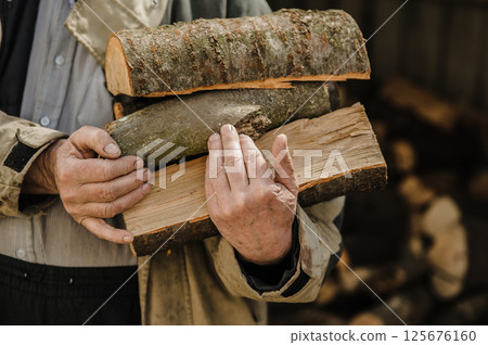Rough, veined hands carry freshly chopped logs. Behind them, firewood is piled high. 125676160