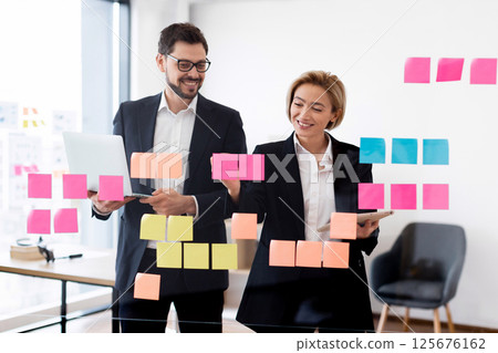 Business colleagues, young man and woman, collaborating on project ideas, using colorful sticky notes on transparent board while smiling in office environment 125676162