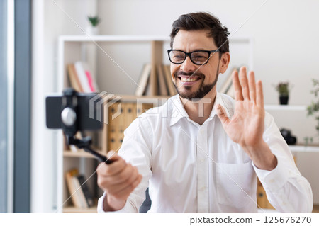 Middle-aged man recording video call with smartphone indoors expressing happiness and engagement. Dressed in white shirt, captured in bright modern office environment. 125676270