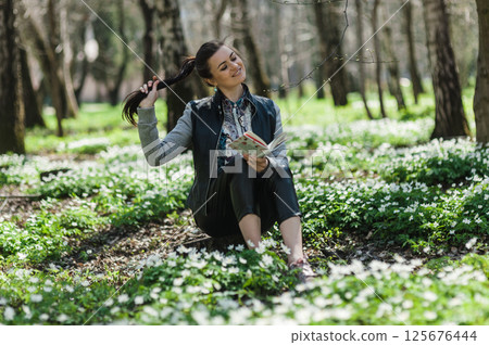 A young girl with a book in her hands is fixing her hair and sitting on the green grass in the forest. A woman with a book is sitting on green grass. 125676444
