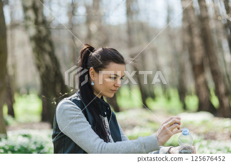 A girl holding a plastic bottle of water in her hands and looking at it in the park. Beautiful woman in the park with a bottle of mineral water in her hands 125676452