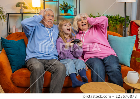 Child girl granddaughter grandparents using joystick controller playing console videogame at home Child girl granddaughter grandparents using joystick controller playing console videogame at home 125676878