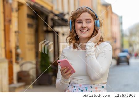 Happy young woman in wireless headphones choosing, listening music in smartphone dancing outdoors 125676905