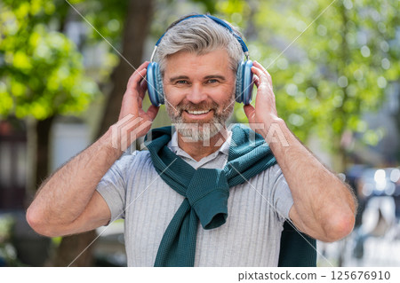 Happy mature man in wireless headphones choosing, listening music in smartphone application outdoors 125676910